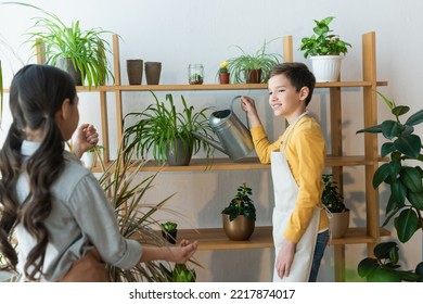 Smiling Boy Watering Plant Near Blurred Friend At Home