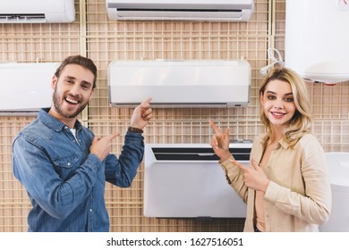 smiling boyfriend and girlfriend pointing with fingers at air conditioner in home appliance store