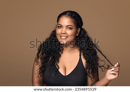 Smiling brazilian body positive lady flirting and touching her beautiful curly hair, looking and smiling at camera over brown background, copy space