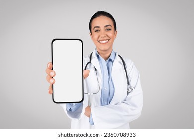 Smiling brazilian female doctor showing smartphone with blank screen at camera, standing isolated on grey background, mockup. Latin therapist woman recommending online medical offer - Powered by Shutterstock