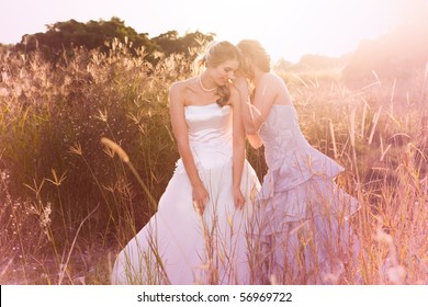 A smiling bride wearing a white wedding dress is listening to her bridesmaid in a rural landscape tell her a secret. Horizontal shot. - Powered by Shutterstock