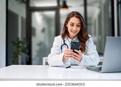 A smiling brunette female doctor texts messages on a mobile phone while sitting in front of the laptop at the office. - Powered by Shutterstock