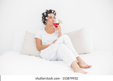 Smiling Brunette In Hair Curlers Sitting On Her Bed Drinking A Cocktail In Bedroom At Home