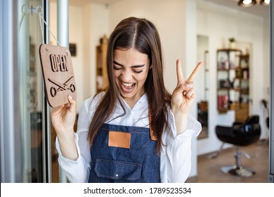 Smiling brunette owner of salon standing with sign open. Smiling owner of hair salon standing with sign open and leaning on glass door. Young female business owner standing outside her salon shop