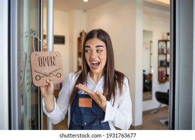 Smiling brunette owner of salon standing with sign open. Smiling owner of hair salon standing with sign open and leaning on glass door. Young female business owner standing outside her salon shop - Shutterstock ID 1931763677
