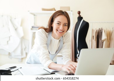 Smiling brunette seamstress in blue apron working with laptop in workshop - Powered by Shutterstock