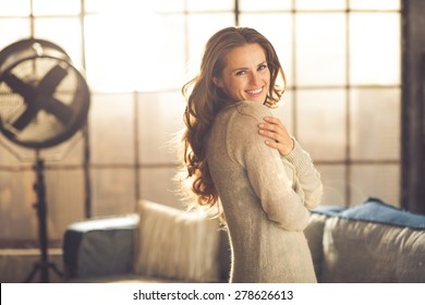 A smiling brunette woman in comfortable clothing is standing in a loft living room, hugging herself while looking over her shoulder. Urban chic loft decoration details.