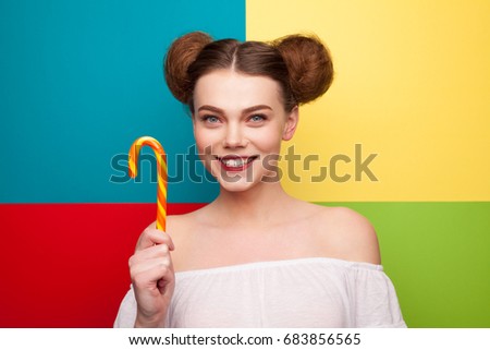 Smiling brunette woman with hairbuns wearing white dress holding candy cane standing against colorful background. 