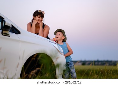 Smiling Brunette Woman Leaning On Hood Of White Car And Looking Down Playfully At Son Outdoors In Green Field At Sunset