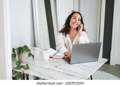 Smiling Brunette Woman With Long Hair In White Cardigan Working On Laptop Using Mobile Phone In Bright Modern Office