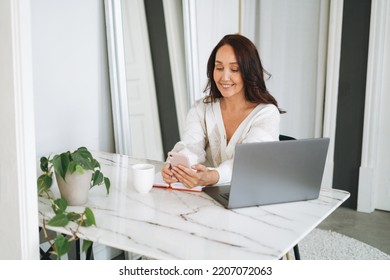 Smiling Brunette Woman With Long Hair In White Cardigan Working On Laptop Using Mobile Phone In Bright Modern Office