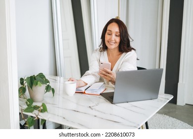 Smiling Brunette Woman With Long Hair In White Cardigan Working On Laptop Using Mobile Phone In Bright Modern Office