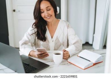 Smiling Brunette Woman With Long Hair In White Cardigan Working On Laptop Using Mobile Phone In Bright Modern Office