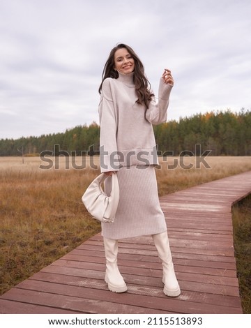 Smiling brunette woman in warm oversize sweater and skirt holds white handbag standing on wooden footbridge at countryside in autumn
