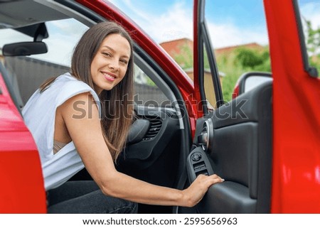 A smiling brunette woman wearing a white sleeveless top sits inside a red car with the door open, appearing happy and ready for a journey or road trip.