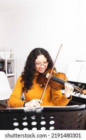 Smiling Brunette Woman Wearing A Yellow Sweater, And Playing Violin By Reading Sheet Music.