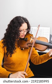 Smiling Brunette Woman Wearing A Yellow Sweater, And Playing Violin By Reading Sheet Music.