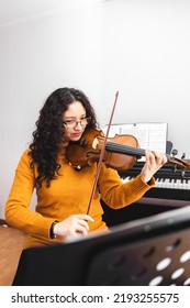 Smiling Brunette Woman Wearing A Yellow Sweater, And Playing Violin By Reading Sheet Music.