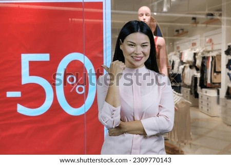 Smiling brunette young asian woman pointing back with her thumb. Boutique with fifty percent discount.