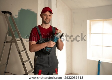 Smiling builder holding an angle grinder during a house renovation with a metal ladder leaning against a partially painted wall and a window allowing natural light to illuminate the scene