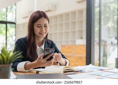 Smiling Business Asian Woman Using Phone In Office. Small Business Entrepreneur Looking At Her Mobile Phone And Smiling.