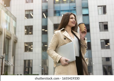 Smiling Business Lady Holding Laptop Under Arm Walking Down The Street Near Office Center With Cup Of Coffee Looking Pleased, Eating On The Go Concept