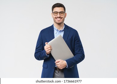 Smiling Business Man Looking At Camera Through Trendy Glasses, Holding Closed Laptop Next To Chest As If Going To Office, Isolated On Gray Background