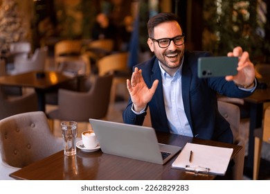 Smiling business man sitting at table in modern city Cafe or restaurant in the evening, waving at mobile phone camera, greeting someone, using smart phone to communicate online, make a video call. - Powered by Shutterstock