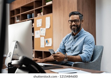 Smiling Business Man Wearing Eyeglasses Working On Desktop Computer While Using Phone In Office. Middle Eastern Businessman Using Smartphone While Sitting At Desk In Office Designer.