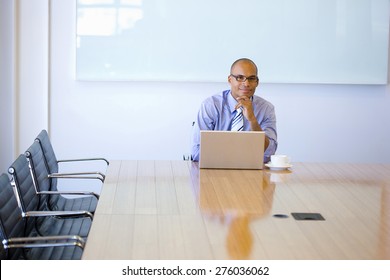 Smiling Business Manager Sitting At The End Of A Meeting Table, With Cup Of Cafe And Laptop.