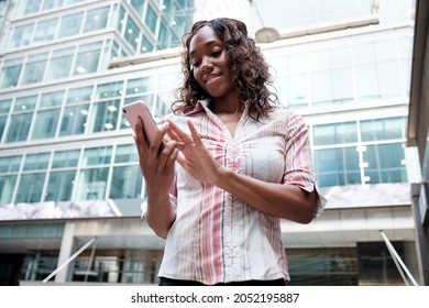 Smiling Business Mature Black Woman Using Smartphone. She Is Wearing A White And Pink Shirt And There Are Business Building Behind.