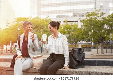 Smiling business partners having break outdoor sitting in a square. Happy mature woman and her colleague drinking coffee in city centre. Two smiling women in conversation outside office, copy space.