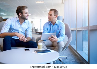 Smiling Business Partners Sitting In A Bright Modern Office Space, Talking Positively And Using A Digital Tablet To Share Information