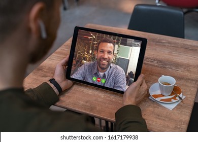 Smiling Business Partners Talking Through Video Call. Back View Of Man In Coffe Shop Doing Video Chat Calling A Friend. Young Man And His Friend Talking To Each Other Through A Videocall On Tablet.