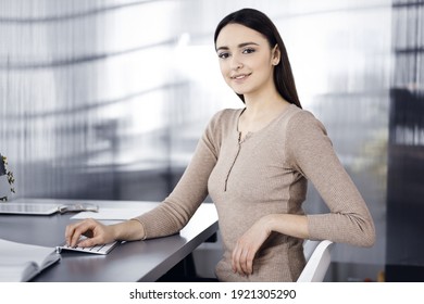 Smiling business woman casual dressed is looking at camera, while sitting at the desk in a modern office. Concept of business success