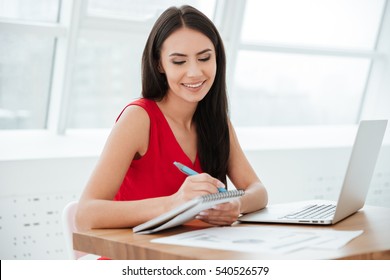 Smiling Business Woman In Red Shirt Sitting By The Table With Laptop And Writing Something In Office