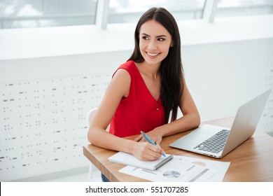 Smiling Business Woman In Red Shirt Sitting And Writing Something By The Table As Well As Looking Away