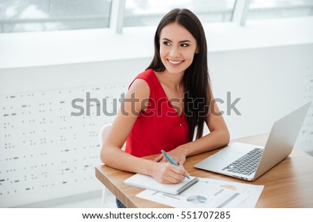 Smiling business woman in red shirt sitting and writing something by the table as well as looking away