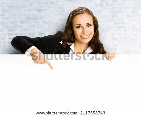 Smiling business woman standing behind, peeping from blank banner or mock up signboard, showing copy space, on white bricks loft wall background. Big board, bill big board, advertisement billboard.