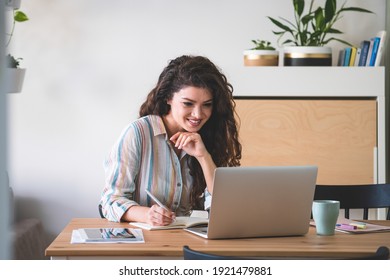 Smiling Business Woman Teleconferencing From Home. Happy Businesswoman Sitting And Talking To Colleagues In A Meeting Using Her Laptop Computer And Writing Down Notes