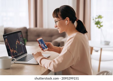 Smiling business woman trader analyst looking at laptop monitor, holding smartphone, wearing earphones. Investor broker analyzing indexes, trading online investment data on stock market graph at home - Shutterstock ID 2095299151