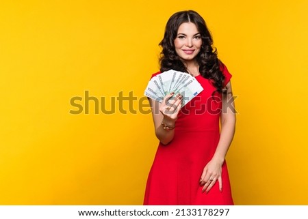 Smiling business woman wears red dress showing fan of cash money in euro banknotes in yellow background in studio. Rich girl looks aside. Business, finance, saving, banking