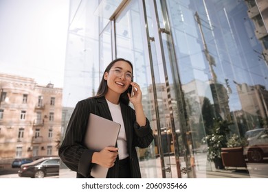 Smiling Business Young Asian Woman Working In Her Luxury Office Holding Laptop At Panoramic Door. Long-haired Brunette In Fashionable Glasses Talking On Phone. Fashion, Technology And People Concept.