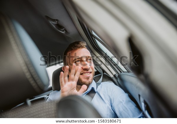 A smiling businessman in the backseat of a car\
looks out the window and\
waves