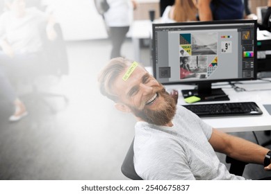 Smiling businessman with a beard in a casual office setting, sitting by a computer. Creative business workspace with design software on screen. Relaxed, modern office vibe. Casual businessman concept. - Powered by Shutterstock