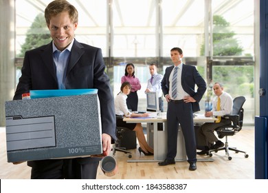 Smiling businessman carrying his things out of the office in a box after quitting his job as his boss and colleagues watch him leave