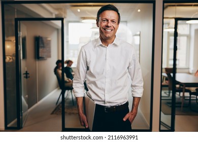 Smiling Businessman Carrying A Laptop Standing In An Office With A Group Of Colleagues Having A Meeting In The Background