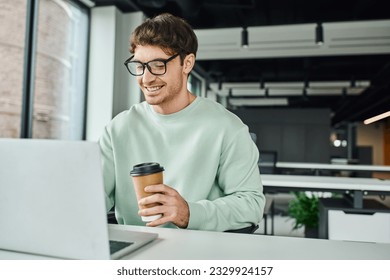 smiling businessman in casual clothes and stylish eyeglasses holding paper cup with takeaway drink and working on laptop in modern office environment, successful business concept - Shutterstock ID 2329924157