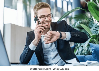 Smiling businessman checking time on watch while working on laptop and talking on smartphone while sitting in a cafe. Entrepreneur looking at wrist with watch waiting for meeting with colleague - Powered by Shutterstock