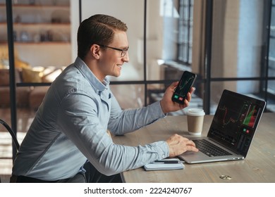 Smiling businessman crypto trader holding smartphone checking candlestick graph on laptop, buying and selling currency, happy to notice tendency of btc rising up, golden bitcoin near on table - Powered by Shutterstock